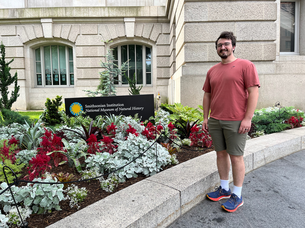 A person stands next to a garden. A building is visible in the immediate background. 