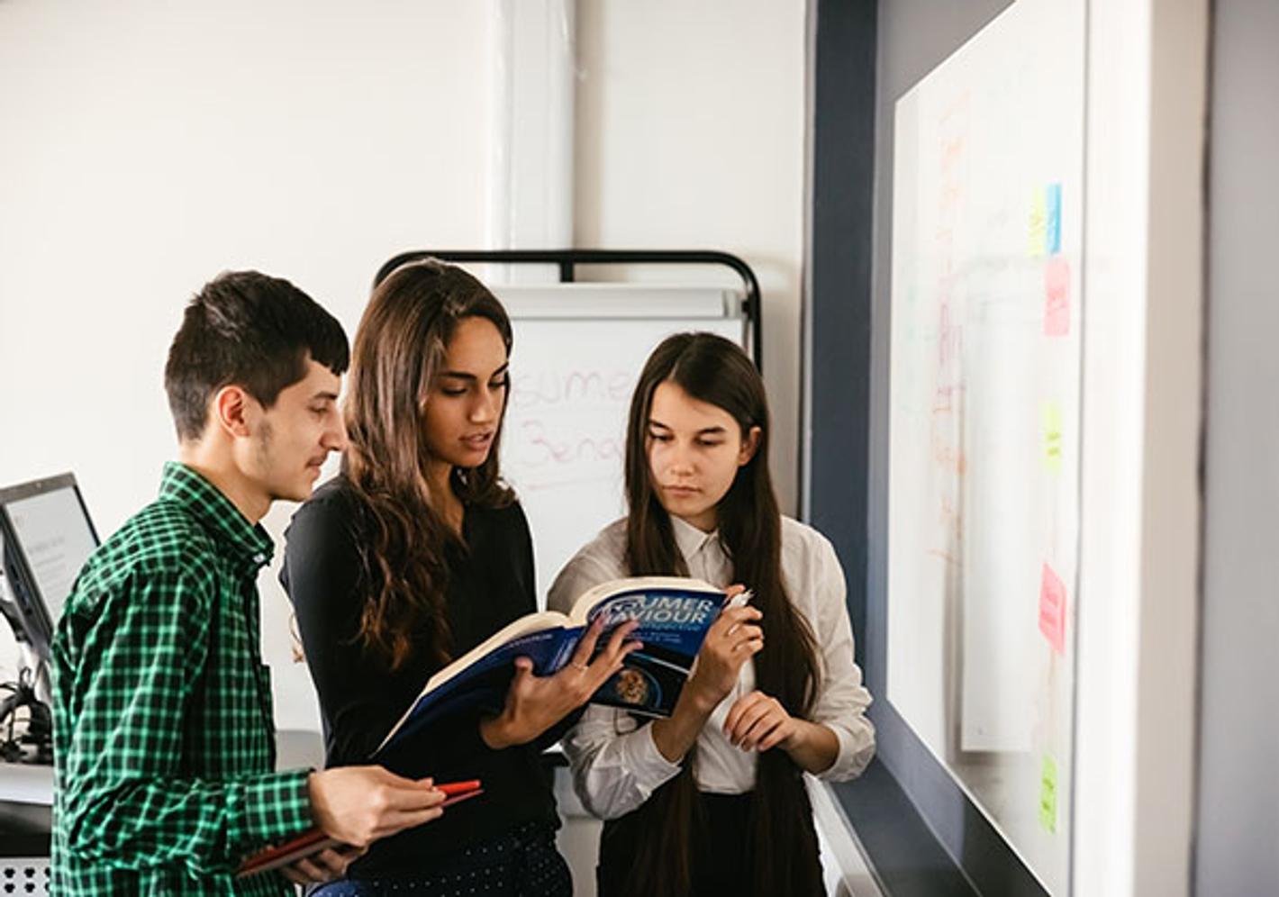 three students study book in front of white board