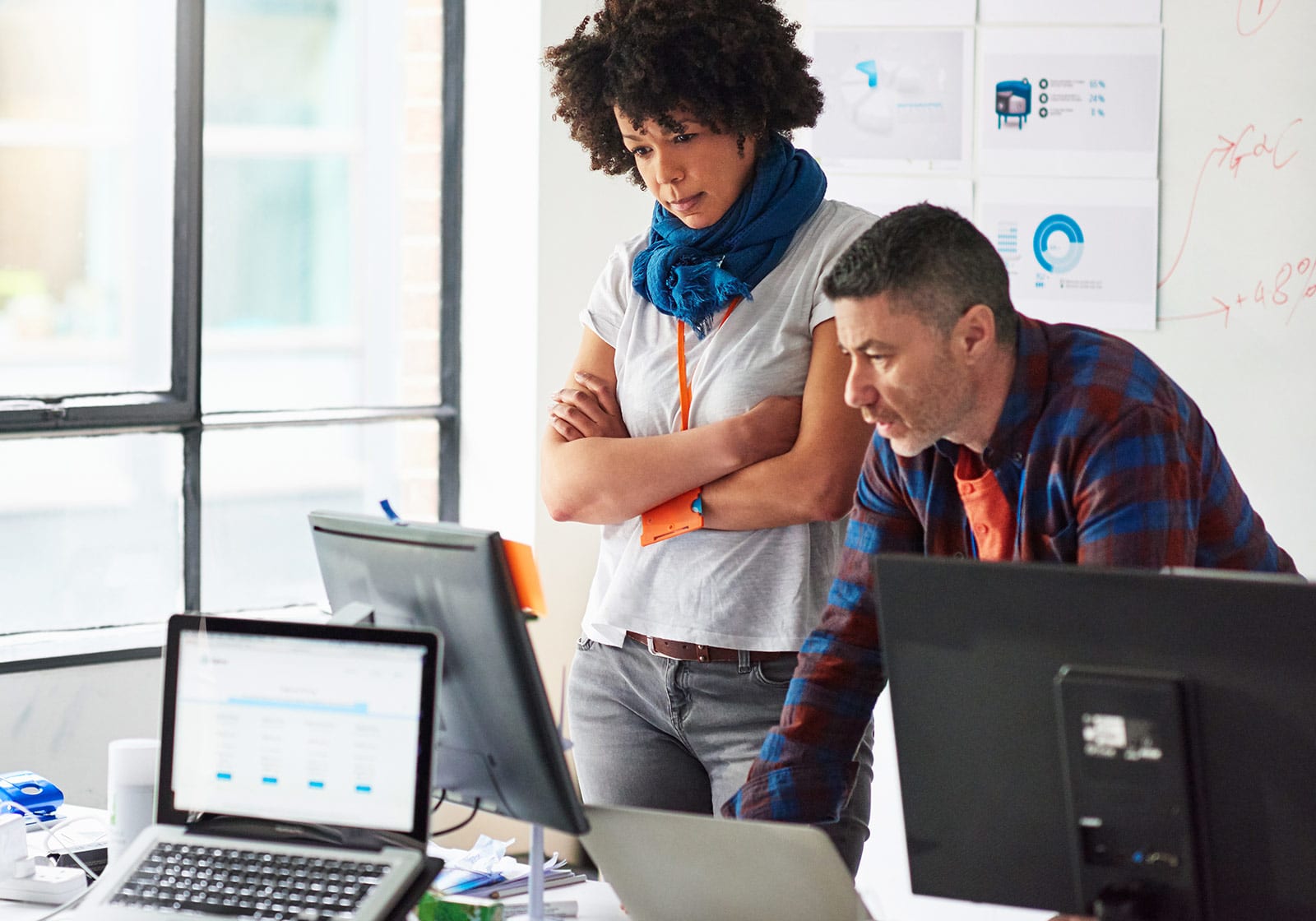 Two people are working at a desk with computers and office supplies, looking intently at a computer screen in a well-lit office space. Charts and diagrams are visible on a wall in the background.