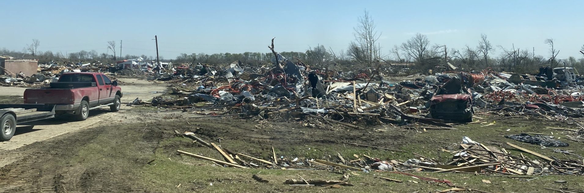 Debris litters the ground, on a sunny day. Where once there was a town with several houses, now there is just splinters and dead trees with no leaves.