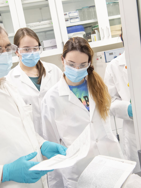 Three people wearing white lab coats, latex gloves, and masks in a chemistry lab