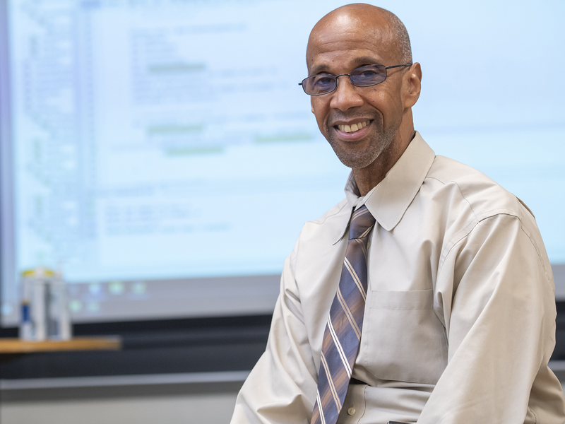 Instructor wearing button down shirt and tie sits in front of a projector with coding information on screen