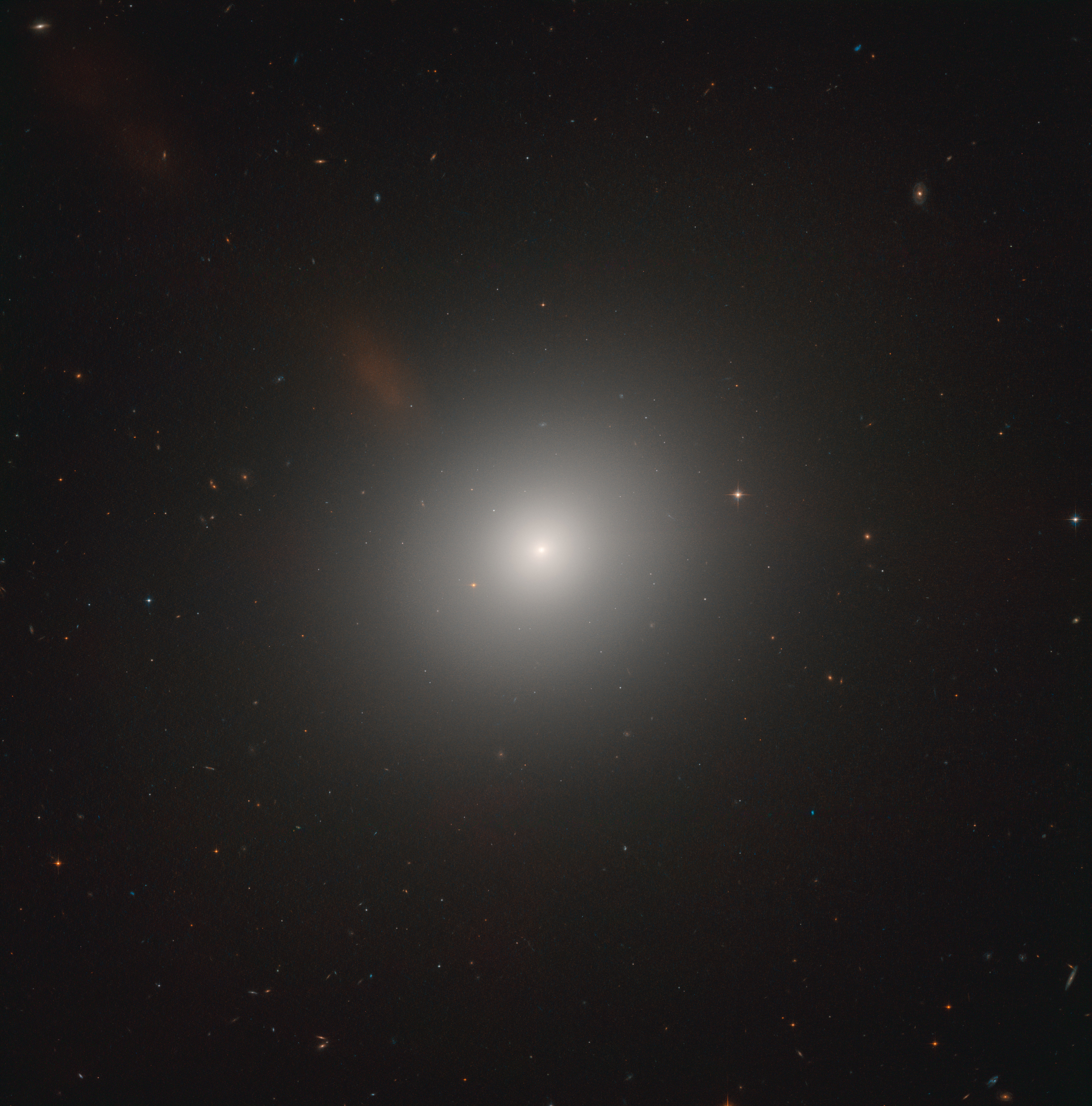 The bright-white, diffuse glow of an elliptical galaxy sits at image center. The galaxy's core appears as an intense-white circle that gets more diffuse as you move outward from the core. A rusty-red, diffuse cloud is visible to the upper-left of the galaxy. It extends to the upper-left corner of the image, where it is very faint. Black background dotted with foreground stars and distant galaxies.