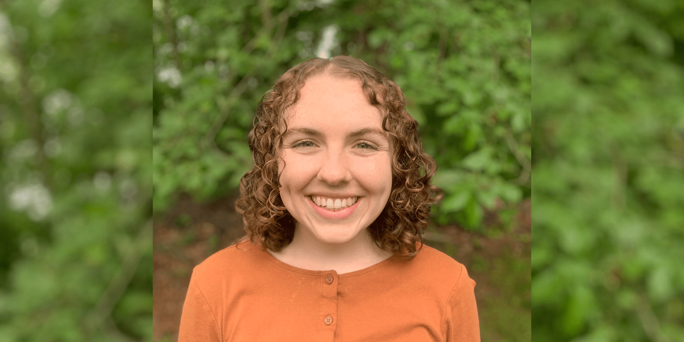 Student with red curly hair and orange top stands in front of greenery and smiles at the camera