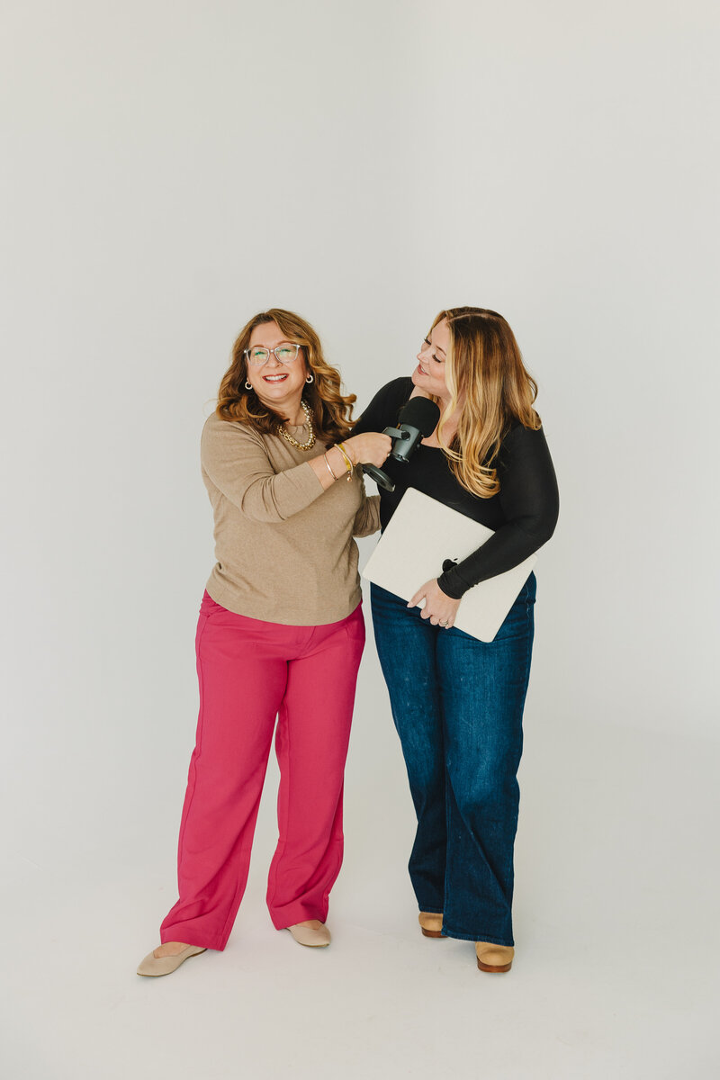 cory and melissa on a white background. melissa holding a laptop and corry holding a micrphone out to pretend to interview her