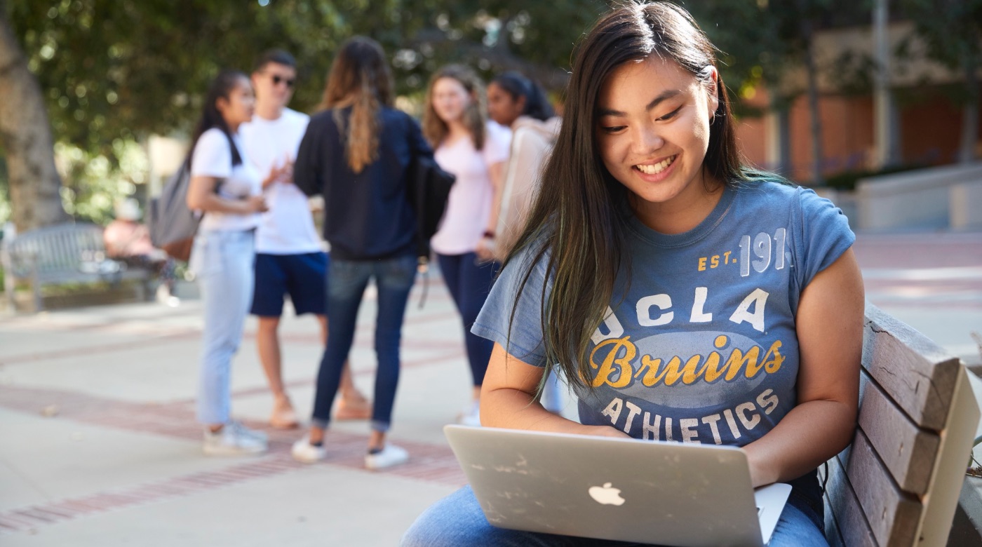 A student sitting on a bench and working on her laptop as a group of five students behind her have a conversation.