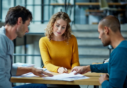 Two men and a woman work together on some papers in the middle of the table in an office environment.