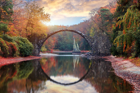 Fantastic Autumn Landscape. Amazing sunset With colorful sky in Azalea and Rhododendron Park Kromlau .Rakotz Bridge, Rakotzbrucke Devil's Bridge in Kromlau, Saxony, Germany.