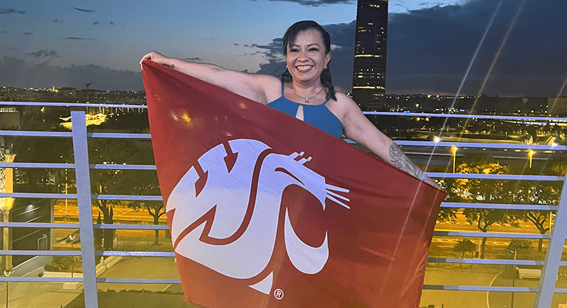 Student Abi Jaimes holding a crimson WSU flag with the Coug head on it with a night cityscape behind her.