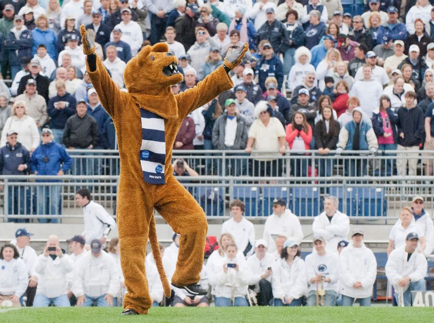 Nittany Lion mascot standing on football turf with arms raised in front of Penn State football fans mainly wearing white and blue.