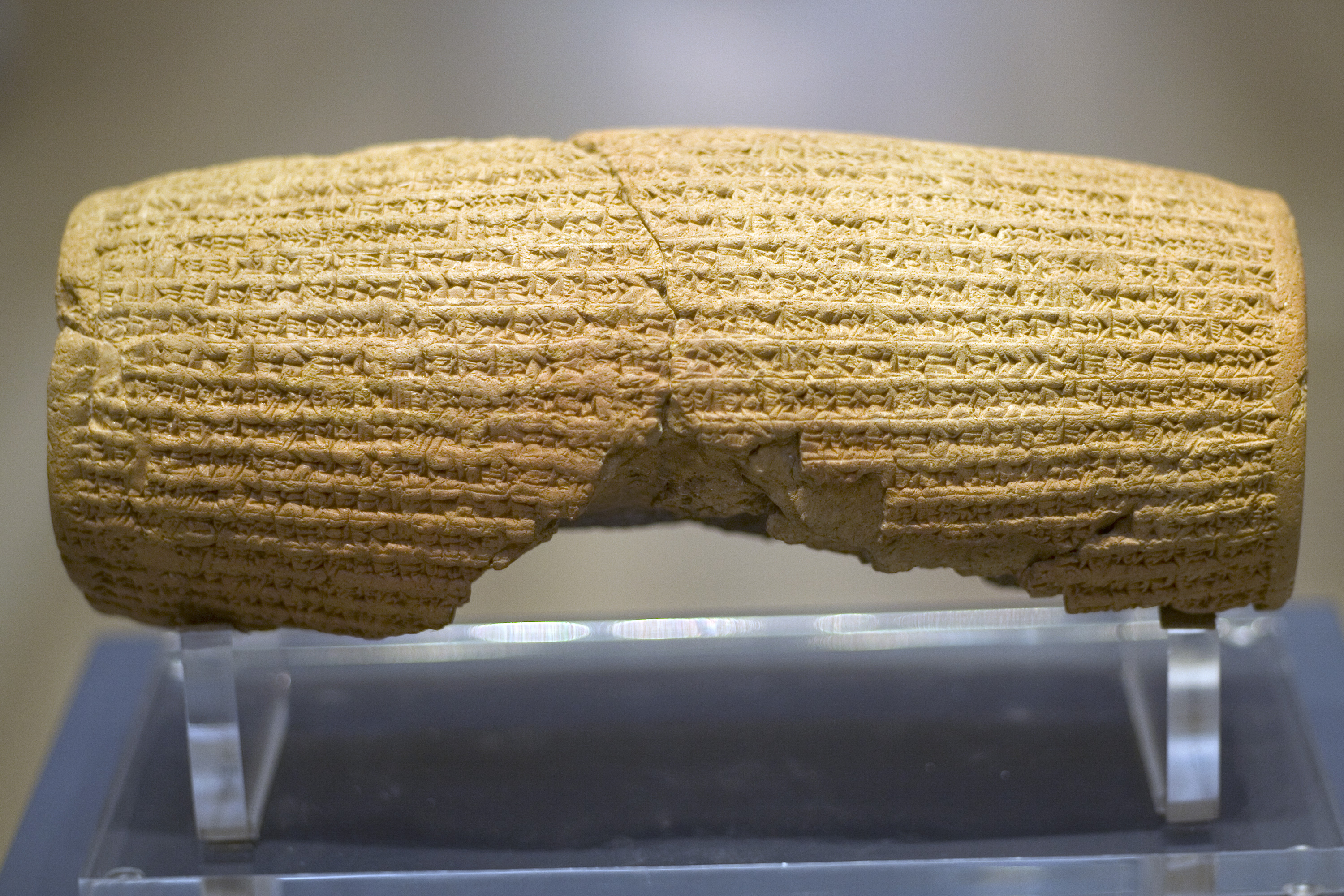 Rear view of a barrel-shaped clay cylinder resting on a stand. The cylinder is round with square edge very close up. It is covered with lines of cuneiform and has a hollow centre. The centre is lined with plate measuring around 10 millimeters in thickness.text