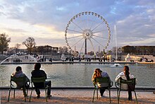 Frontale Farbfotografie eines Riesenrads mit zwei steigenden Wegen und zwei Statuen an beiden Seiten. Um das Riesenrad sind Buden und hinter ihm steht ein Obelisk. Im Vordergrund ist eine runde Wasserfläche mit vier Fontänen und um ihr herum sind Stühle und Menschen. Am unteren Bildrand sitzen paarweise zwei Männer und zwei Frauen mit dem Rücken zur Kamera auf Stühlen.