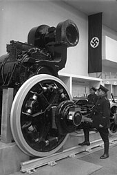 A traction motor from a German E18 class at an industrial exhibition. There is a Nazi swastika flag hanging behind and a man in military uniform is striking a heroic pose as if admiring the motor.