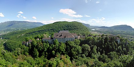 Vue des bâtiments d'un château sur les pentes boisées d'un éperon rocheux.