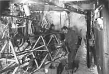 Photographie en noir et blanc de l'intérieur d'un hangar. Plusieurs hommes habillés chaudement s'affairent autour d'un structure métallique.