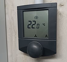 Plochý černý přístroj umístěný na stěně. Na černobílém displeji přístroje je zobrazena hodnota 22,0 °C a ikona panáčka v domečku.