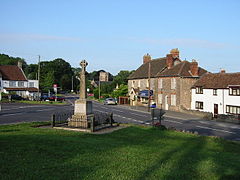 Stone cross surrounded by railings on grass area in front of roads and houses.