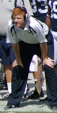 Standing on a football field sideline, leaning forward with hands on his spread-apart legs above the knees; McQueary has red hair and is wearing headphones, a white shirt, and dark pants.