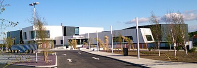 Sports centre building, with white and black cladding, set in large car park with landscaping