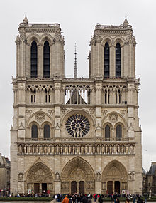 Frontale Farbfotografie einer hellen, gotischen Kathedrale mit zwei breiten, eckigen Türmen und vielen Ornamenten. Alle drei Eingänge haben Spitzbögen mit mehreren Figurenreihen, in deren Mitte Szenen dargestellt sind. Darüber sind eine Figurenreihe, eine Rosette und vier kleine Rundbogenfenster. Die oberen Glockentürme haben lange, schmale Öffnungen. Im Hintergrund ist eine schmale Turmspitze zu sehen.