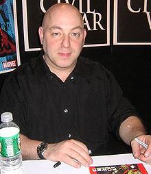 Man with shaved head sits at a table, a botte of water beside him. He holds a pen, and is looking into the camera.