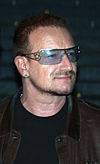 A man with facial hair wearing a leather jacket, a black shirt, an earring, and tinted glasses with a star along the frame..