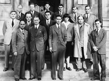A group photo of 14 men in suits and 1 woman in three rows facing the vewier.