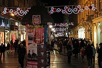 Ulica nocą, z budynkami i sklepami po obu stronach oraz spacerujący tłum ludzi, a ponad nimi świąteczne światła zawieszone pomiędzy budynkami.