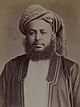 A black-and-white photograph of a man with a dark beard wearing a turban, a dark jacket, a shirt, and a belt, sitting in a chair, and looking at the viewer