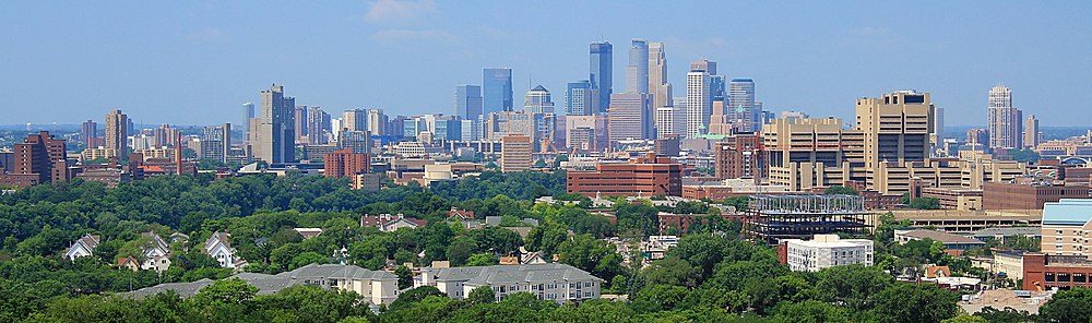 The Minneapolis skyline rises to its highest point at the center of the image, with the three tallest buildings standing out against a clear blue sky. Before the skyline are trees, university buildings, and residential complexes.