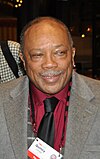 An older man with a white mustache, wearing a black tie, red dress shirt, gray jacket, and a name badge that hangs from a string around his neck.