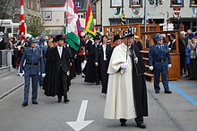 Photographie d'un groupe de personnes en costume sombre, encadrés par deux gendarmes en uniforme bleu ; devant, un homme en costume noir et blanc portant un sceptre ; derrière eux, plusieurs drapeaux portés.