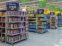 Aisles in a supermarket with small sets of shelves on the end, receding from left to right. Atop each set of shelves is a large white on black sign with the price of an item for sale on those shelves. White on green signs with the types of products in the aisles hang from the high ceiling.