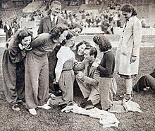 Agenouillé au sol, entouré de femmes, un homme écrit sur les vêtements de l'une d'entre elles avec un stylo.