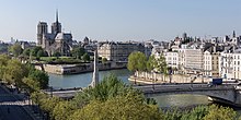 Farbfotografie in der Obersicht von zwei Stadtinseln mit vielen eng anliegenden Altbauten. Der Fluss verläuft von unten rechts und teilt sich in der Mitte auf. Im Vordergrund ist eine Brücke mit Autos und Fußgängern, die links unten in eine Allee und eine hohe, weiße Statue führt. Hinter dem linken begrünten Ufer ist eine Kathedrale mit zwei Ecktürmen und einem gotischen Turm.