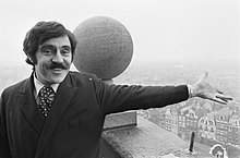 Photo en noir et blanc d'un homme brun moustachu souriant qui désigne du bras une ville qui s'étend en contrebas