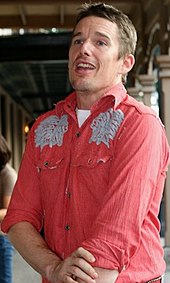 A caucasian male with light brown hair and light brown sttuble is facing to the left. He is wearing a red shirt, with a white t-shirt visible underneath. The red shirt has a white pattern on the shoulders.