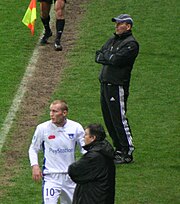 Trois hommes debout au bord d'un terrain de football, dont un footballeur et deux personnes en survêtement.