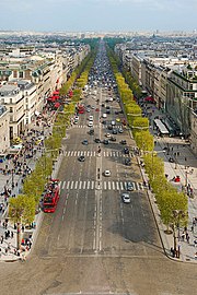 Frontale Farbfotografie in der Obersicht einer zehnspurigen Allee mit viel Verkehr, die bis zum Horizont führt. Vorne links ist ein roter Doppeldecker mit Touristen. An beiden Seiten sind breite Bürgersteige mit vielen Menschen und vielen Häusern. Im Hintergrund ist ein Stadtpanorama.