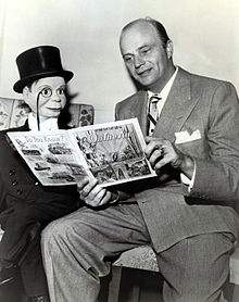 photo noir et blanc d'un homme assis en costume qui semble faire la lecture d'un grand livre à une marionnette également en costume