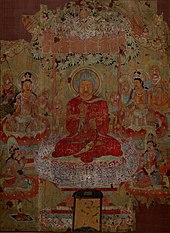 malba buddhistických světců, v centru na ozdobné podušce sedí v pozici lotosového květu (se skříženými nohama) osoba v červeném oděvu, nad ní se klene zdobná stříška, kolem ní na poduškách sedí několik dalších osob