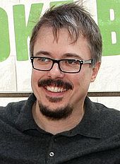 A man with black glasses and a black mustache and beard is smiling at the camera. He is wearing a black shirt.