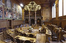 Photographie d'une salle, avec sur la gauche une peinture murale, au centre des pupitres en bois disposés en cercles, au plafond un chandelier.