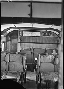 Photograph of the passenger cabin of a Short Kent airliner, showing pullman-style seating and a man sitting in the rearmost row, a map of the Genoa-Alexandria route on the wall.