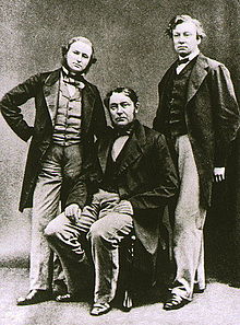 Tiga pria paruh baya, dengan yang di tengah duduk. Semuanya mengenakan jaket panjang, dan pria pendek di sebelah kiri berjanggut.