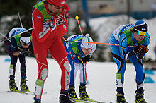 Photographie de quatre hommes faisant du ski de fond, l'un redressé en combinaison rouge, les trois autres penchés vers l'avant.
