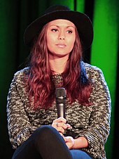 A brunette woman is seated at a chair; she is wearing a hat.