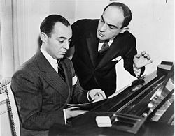 A Caucasian man in his thirties is sitting behind a piano facing left. He has short, dark hair and is wearing a suit jacket, a white shirt and a necktie. He is looking down at a music sheet before him and points to it with his left hand. Another man is standing on his right, also wearing a suit, white shirt and a necktie. He is bent slightly toward the man on the left and looking at him, appearing concentrated in thinking. His eyes are half-closed and his left arm raised as if he was holding a glass.