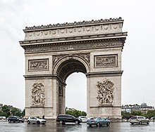 Frontale Farbfotografie eines Triumphbogens mit Autos auf mehrspuriger Straße. An beiden Mauern neben dem Torbogen sind Szenen und Reliefs eingemeißelt. Direkt am oberen Bogenrand befinden sich zwei Figuren mit Posaunen und um die ganze obere Wand sind durchgehend ein Relief und Ornamente zu sehen.