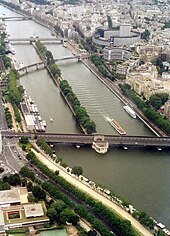 Farbfotografie in der Vogelperspektive von einem Fluss mit einer langen, schmalen Insel, auf der Bäume aufgereiht sind. Die Insel dient als Stütze für drei Brücken, die beide Stadtteile verbinden. Auf der vorderen Brücke fahren Autos und eine Eisenbahnbrücke führt über sie. Unten links und oben rechts sind viele Häuser und Uferalleen mit Schiffen.